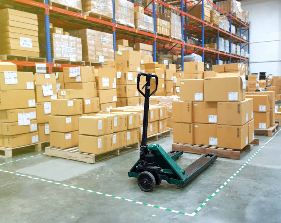 Hand Pallet Truck (Handlift) in industrial warehouse with shelves and cartons on pallets *Barcode and label on cartons have been blurred*